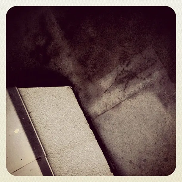 Close-up of a dimly lit corner with intersecting concrete surfaces, textured stains, and a white panel in the foreground.
