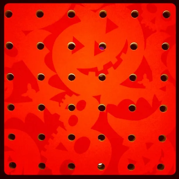 Bright orange pegboard featuring a repeating pattern of jack-o’-lantern faces in darker orange tones, with evenly spaced circular holes.