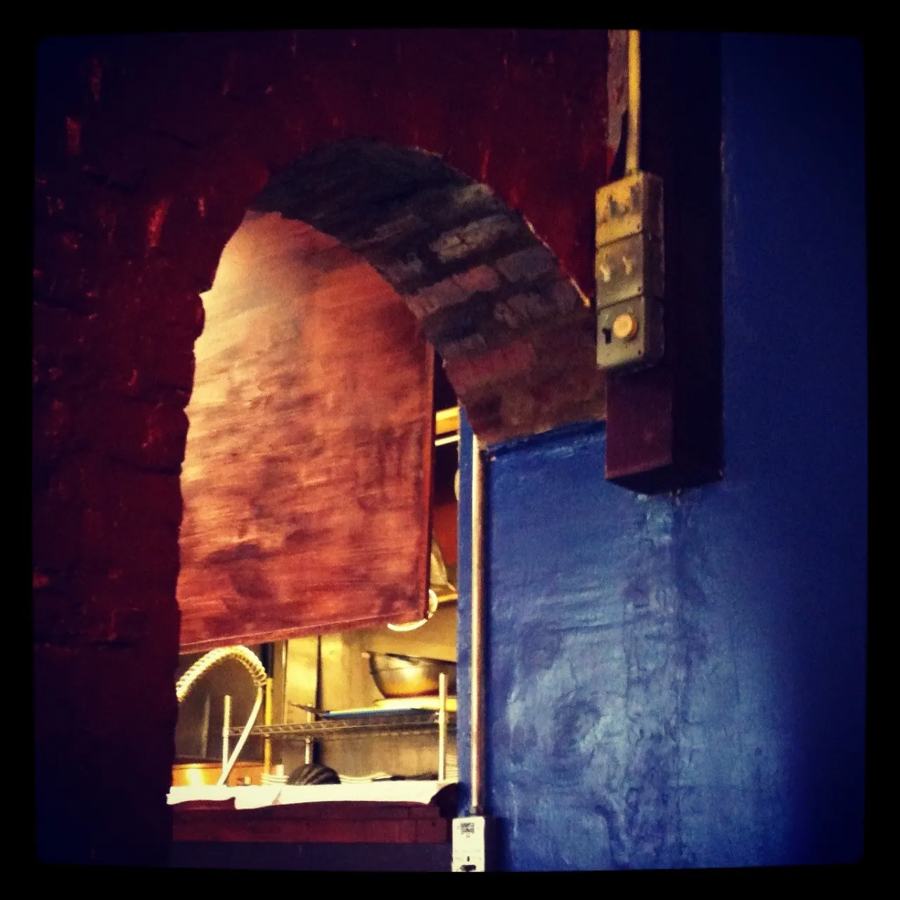 Interior scene showing a red brick archway and blue-painted wall with a vintage doorbell button, leading into a kitchen with visible cookware.