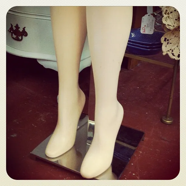 A pair of mannequin legs mounted on a metal base in a store, with furniture, price tags, and a tiled floor in the background.