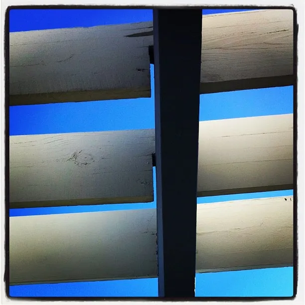 Wooden slats painted white are arranged horizontally above, viewed from below against a bright blue sky with a dark vertical beam dividing the frame. 