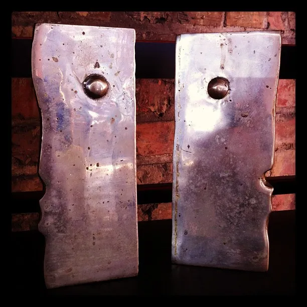 Two abstract metallic sculptures with bolt-like protrusions, standing upright against a brick wall on a dark surface.