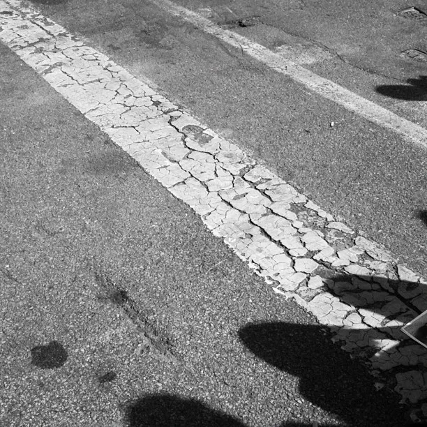 A cracked and weathered white painted line runs diagonally across an asphalt road surface, with visible shadows falling across it.