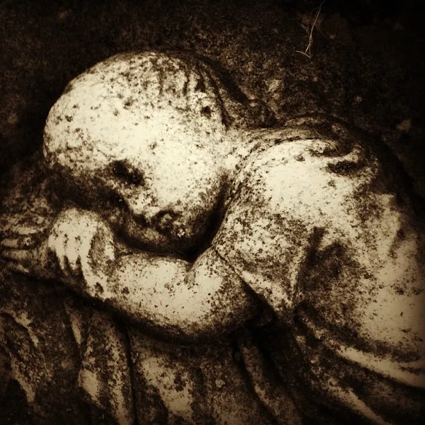Weathered stone sculpture of a child resting its head on folded arms, with a somber expression and textured surface.