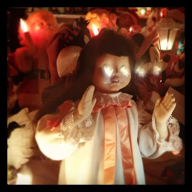 Vintage doll with glowing eyes, dressed in a white gown with lace and ribbon, surrounded by illuminated holiday figures.
