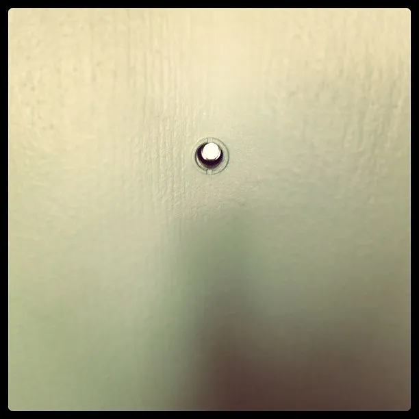 Close-up of a peephole centered on a smooth, light-colored door, with a faint shadow falling vertically beneath it.