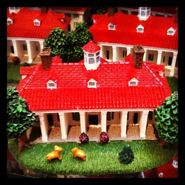 Miniature model of a classical building with red roof, white columns, green trees, and two small orange animal figurines on the lawn.