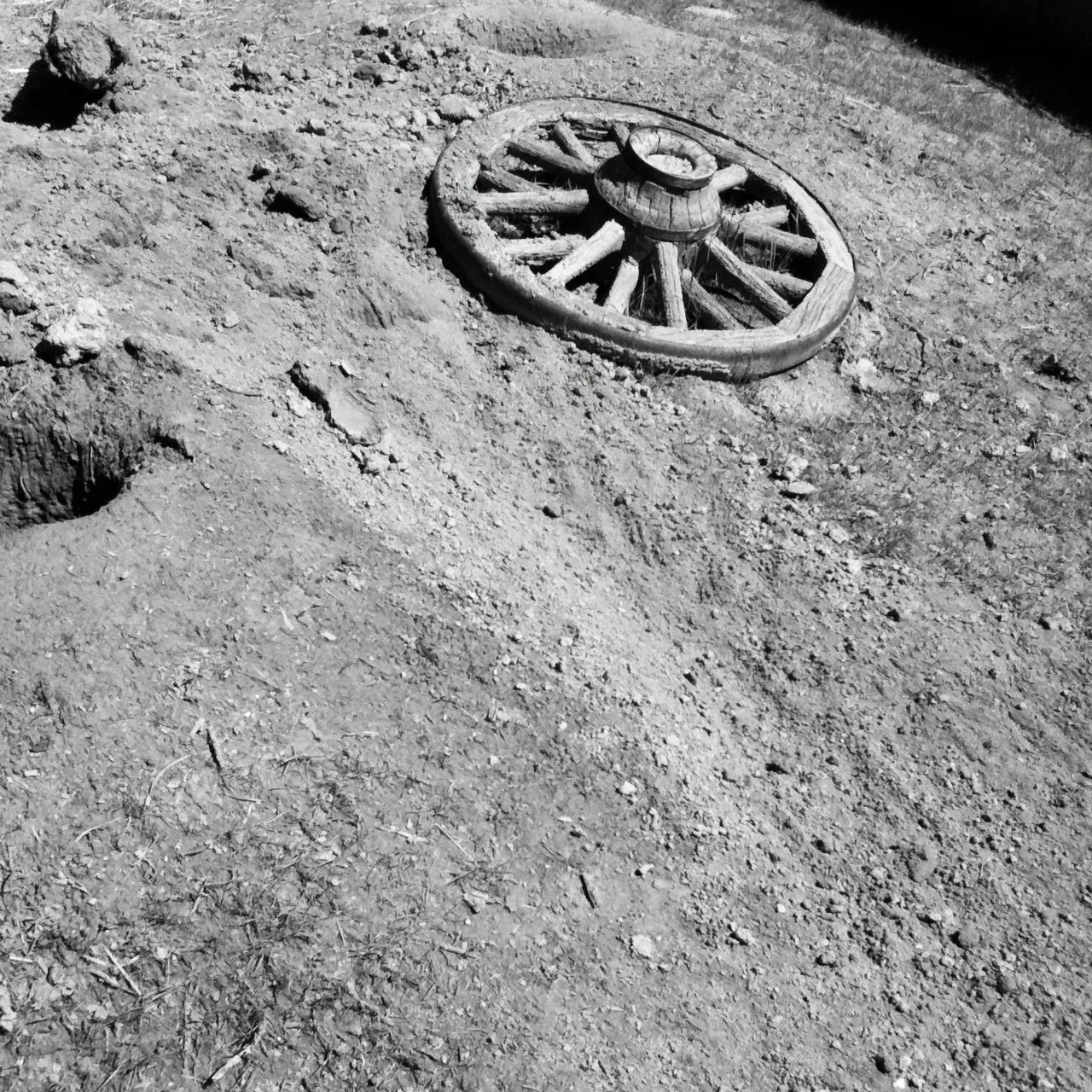 Black and white image of an old wooden wagon wheel partially buried in dry, uneven dirt with scattered rocks and sparse grass.