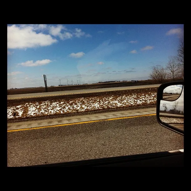 View from a car window of a highway with a snowy median, open farmland, and distant wind turbines under a partly cloudy sky.