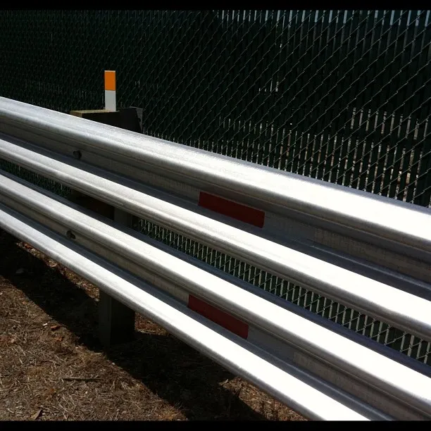 Close-up of a shiny metal guardrail with red reflectors, set against a green chain-link fence and sunlit ground.