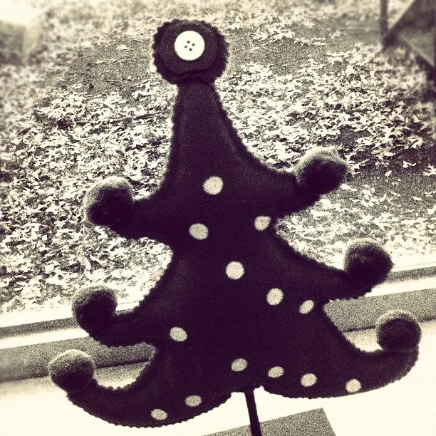 Stylized felt Christmas tree decoration with curled branches and polka dots, topped with a button, positioned by a window with fallen leaves outside.