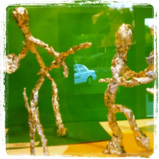 Two humanoid figures made from crumpled foil posed in front of a green background, with a reflection of a white car in glass.