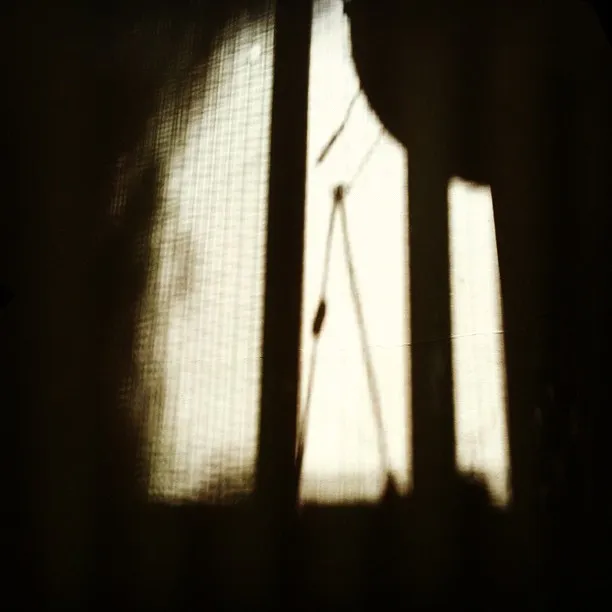 A soft-focus image captures the shadow of a window with cords or blinds cast onto a curtain or screen, creating a moody, abstract composition.