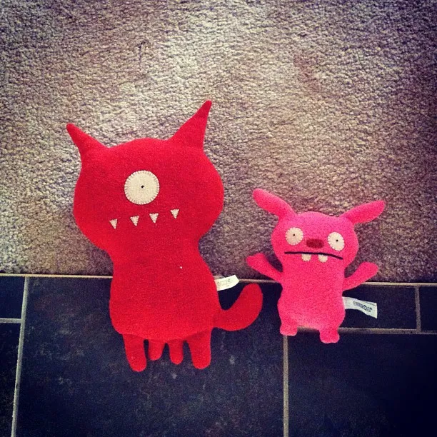 Two plush toys with monster-like designs—one red with a single eye and three teeth, and the other pink with two eyes and a small mouth—are placed on a carpeted floor next to dark tile.