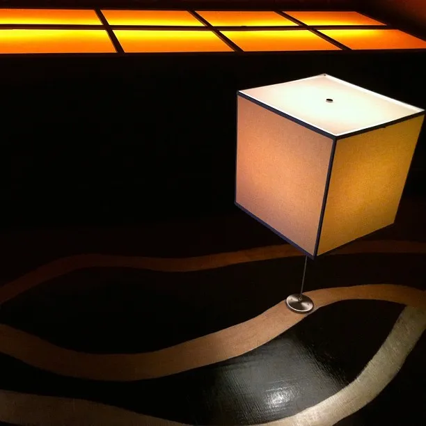 Modern cube-shaped floor lamp on a wavy-patterned dark floor, with a glowing orange grid light on the back wall.