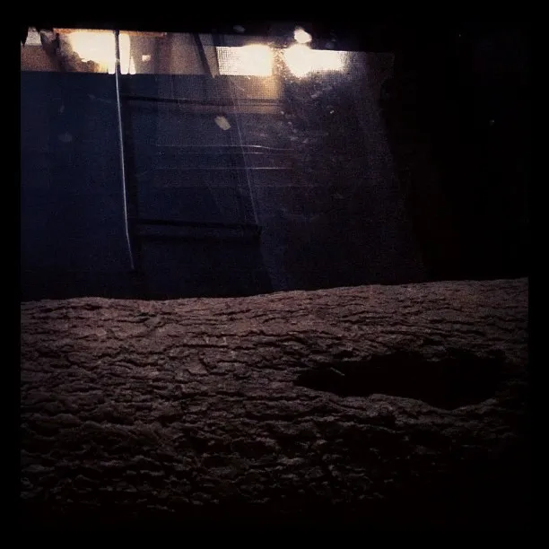A dimly lit scene showing the view through a window with reflections of interior lights and a textured surface in the foreground, possibly a tree trunk or ledge.