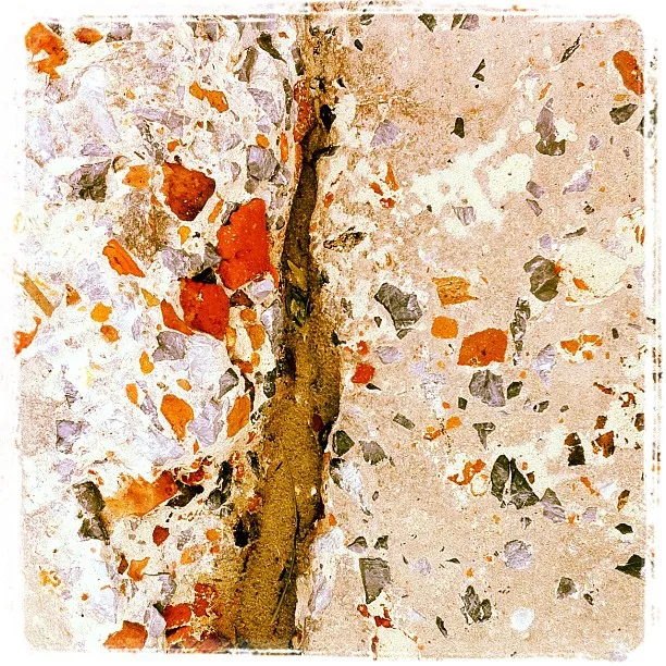 Close-up of a cracked concrete surface with exposed aggregate, showing fragments of red, gray, and white stones.