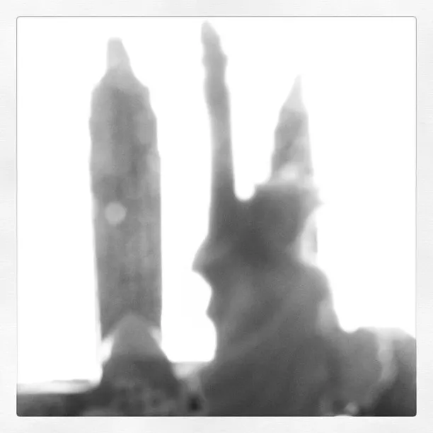 Blurry black and white image of a miniature Statue of Liberty with two tall structures in the background, heavily backlit.