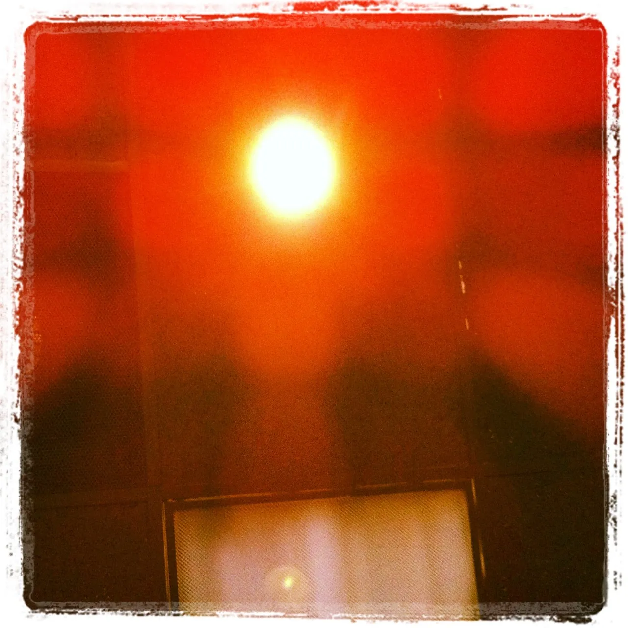 Overexposed ceiling light glowing brightly with intense red lens flare and dark grid ceiling panels in the background.
