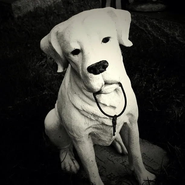 A monochrome photo of a realistic dog statue depicts it sitting on a patch of grass, holding a key on a cord in its mouth, evoking a sense of loyalty or memory.