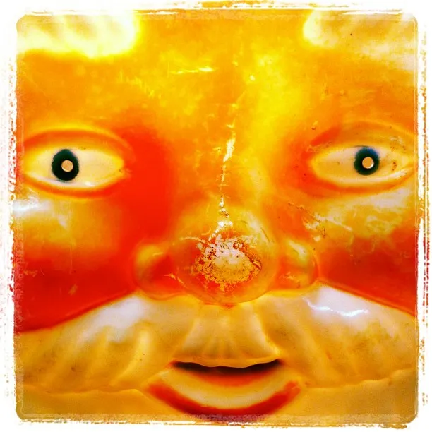 Close-up of a plastic or ceramic sun face with exaggerated features, glowing orange and yellow tones, and a white mustache.