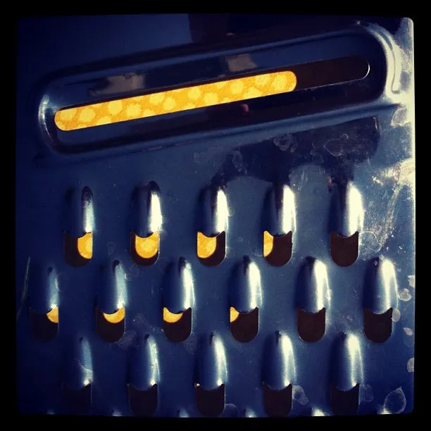 Close-up of a metal cheese grater with a perforated surface and a yellow-orange light source shining through the slots.
