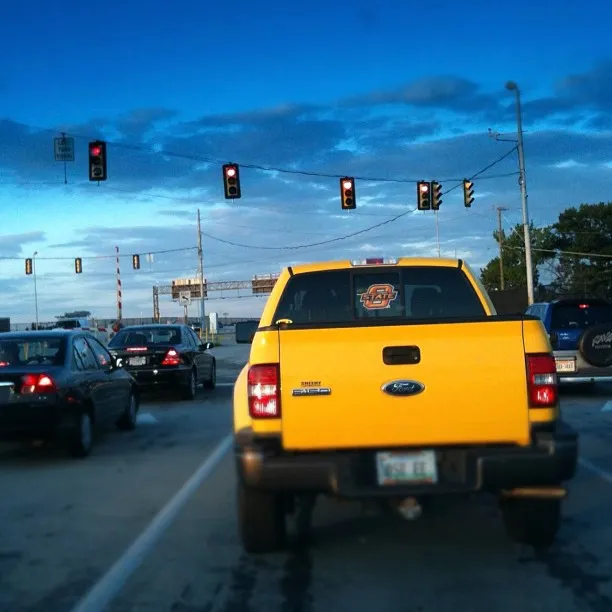 Bright yellow Ford pickup truck stopped at a red light in traffic under a blue sky with scattered clouds.