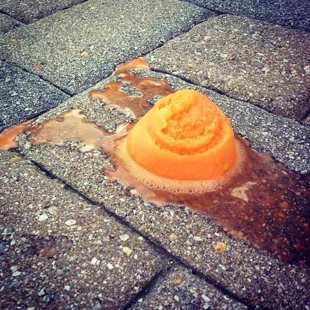 Scoop of orange sherbet melting on textured concrete pavers, with liquid pooling around the base.