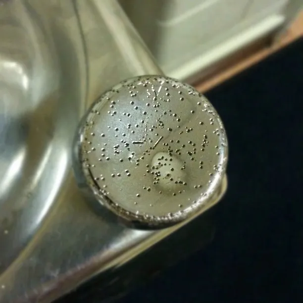 A worn metallic push button on a drinking fountain, with raised Braille markings and signs of wear around the edges.