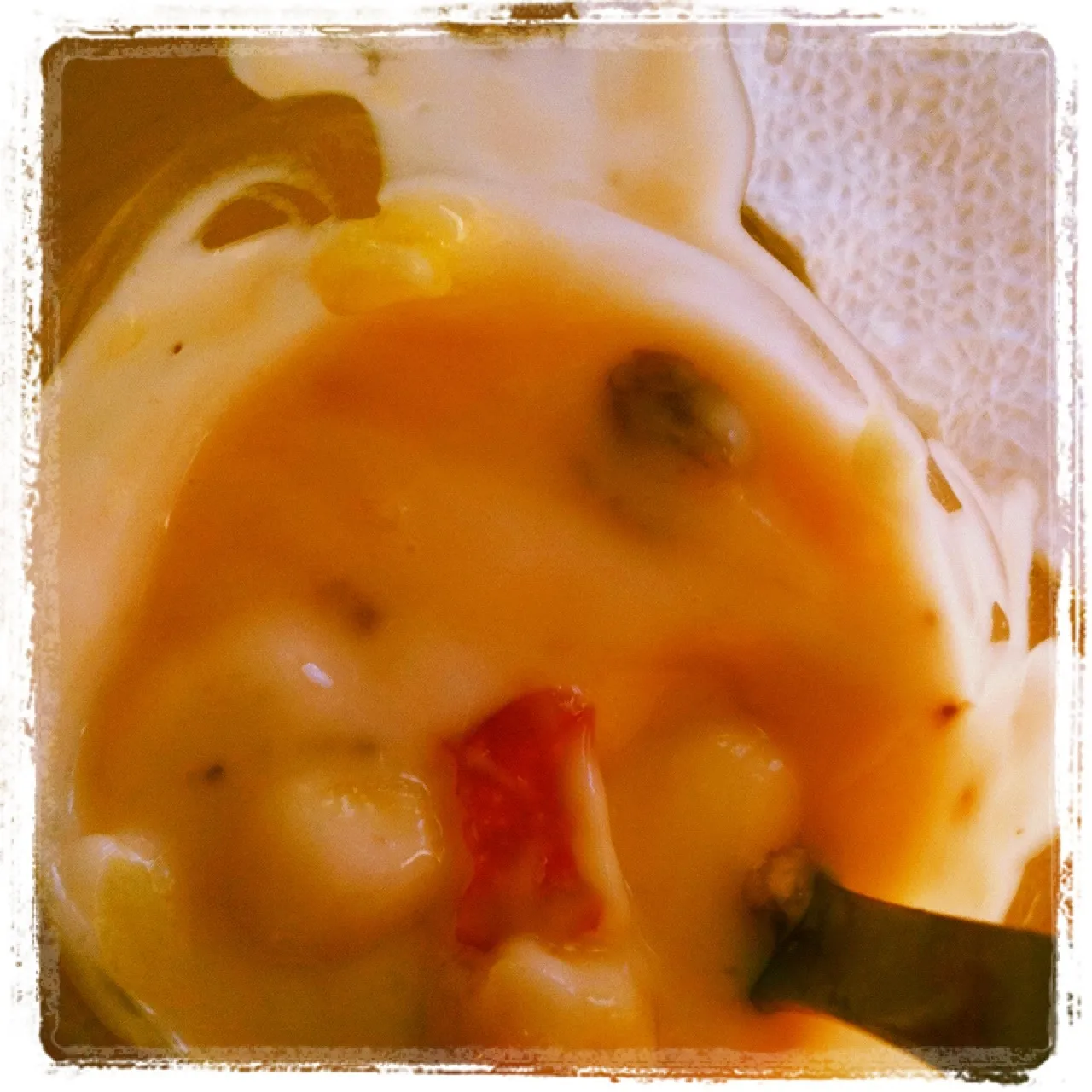 Close-up of a spoon in a creamy fruit yogurt mixture with visible pieces of grape, pineapple, and red fruit in a plastic container.