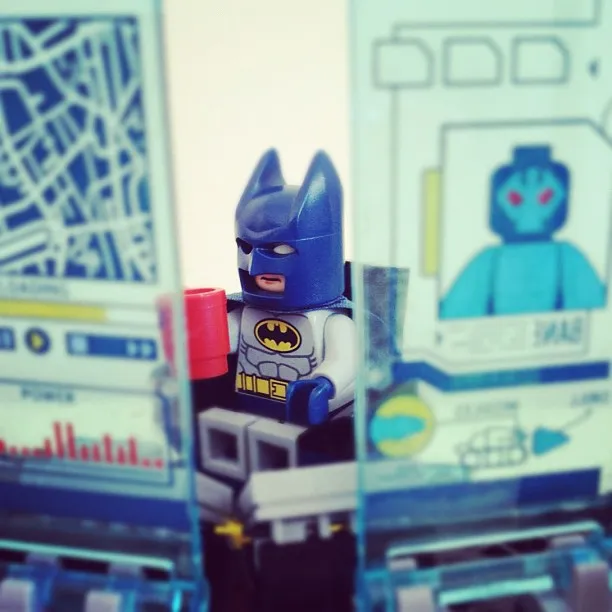 Close-up of a LEGO Batman minifigure sitting between two translucent computer screens with graphics and holding a red mug.