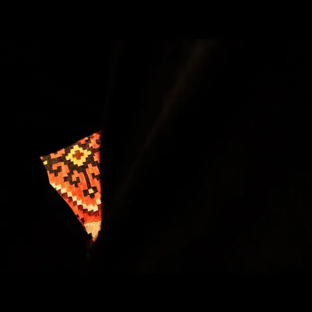 A mostly dark composition with a narrow sliver of patterned orange and black textile visible between deep shadows, suggesting a glimpse through an opening or gap.