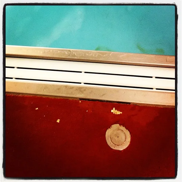Close-up view of the edge of a swimming pool with a metal drain and red flooring, showing a few small crumbs or debris.