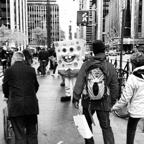 Person in a SpongeBob SquarePants costume standing on a busy city sidewalk among pedestrians in a black-and-white street scene.