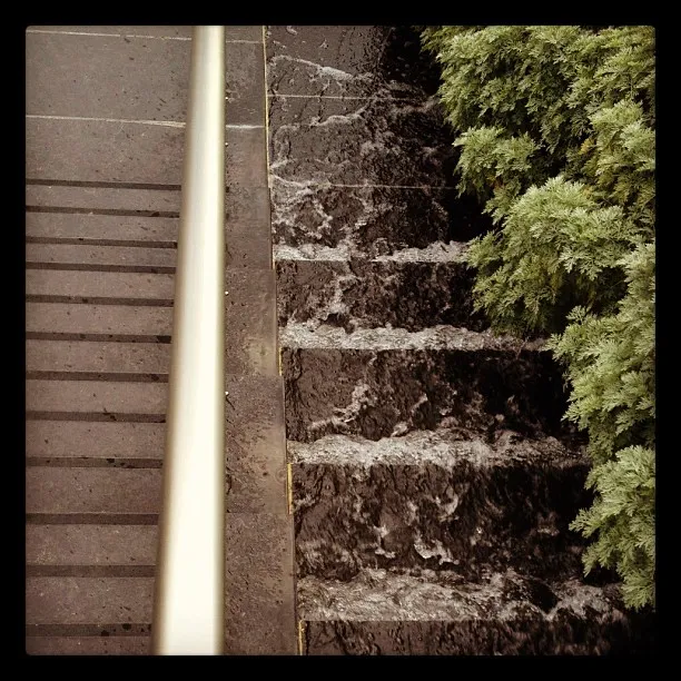 A top-down view of water cascading down dark stone steps beside a metal handrail, with dense green shrubs on the right side.