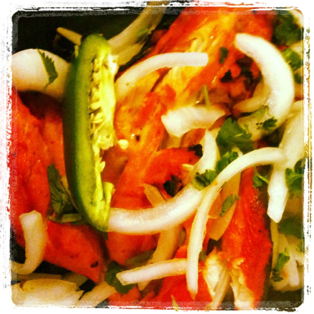 Close-up of a dish with grilled orange-red meat topped with sliced white onions, chopped cilantro, and a halved green jalapeño pepper.