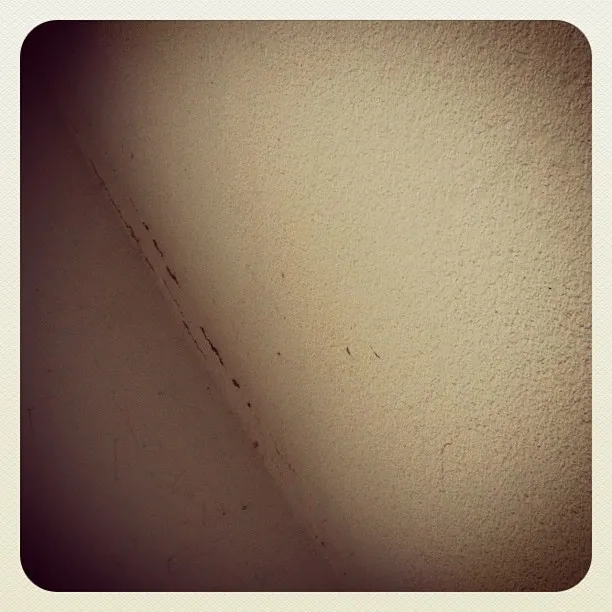 Close-up of a corner where two beige textured walls meet, with visible dirt, cracks, and surface imperfections.