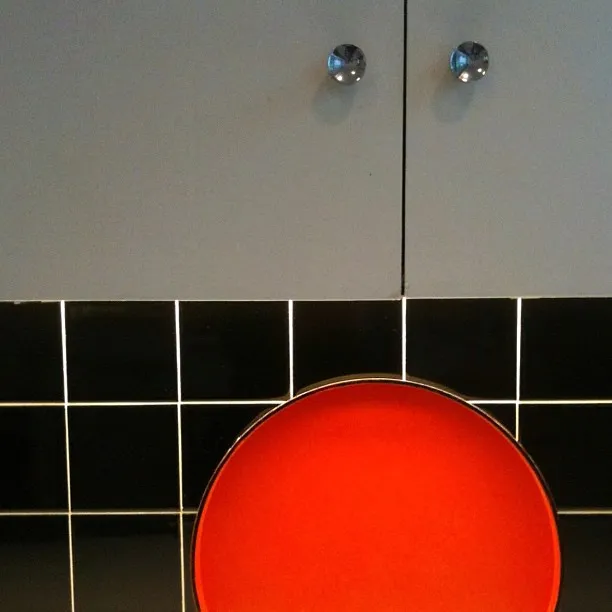 A vivid red circular chair or stool in front of a black tiled wall with white grout, below a cabinet with two crystal knobs.