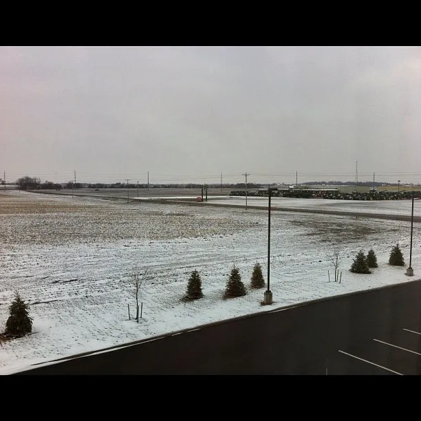 A flat, snow-dusted landscape with sparse trees, empty parking spaces in the foreground, and a gray overcast sky stretching across the horizon.