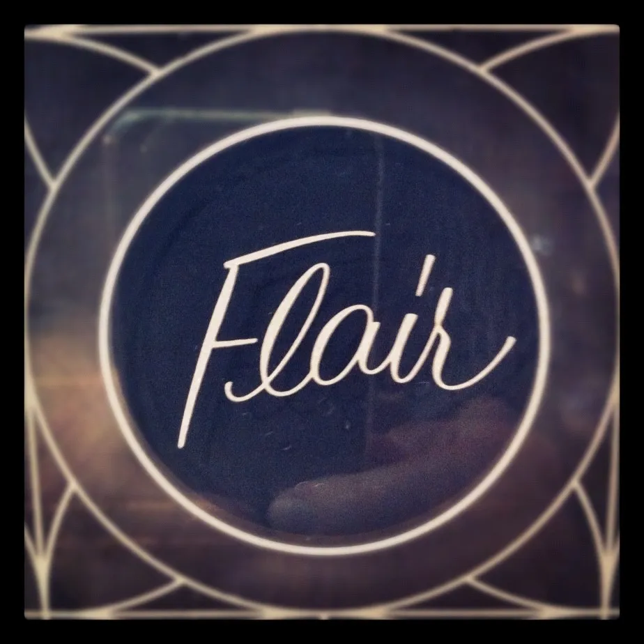 Close-up of a circular logo with the word “Flair” in cursive script, set against a dark background with geometric line patterns.