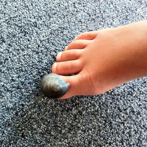 Close-up of a child’s foot with a dark sea snail shell placed on the big toe, resting on a textured gray carpet.