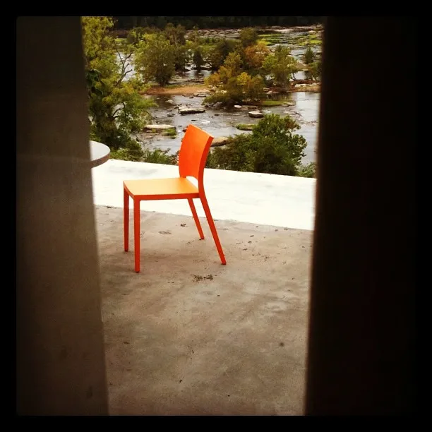 A bright orange chair sits alone on a concrete patio overlooking a river with scattered rocks and lush green trees, partially framed by two vertical elements in the foreground.