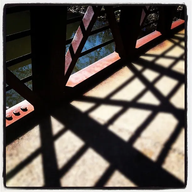 Shadow of a metal bridge railing forming a geometric pattern on a sunlit concrete walkway above a body of water.