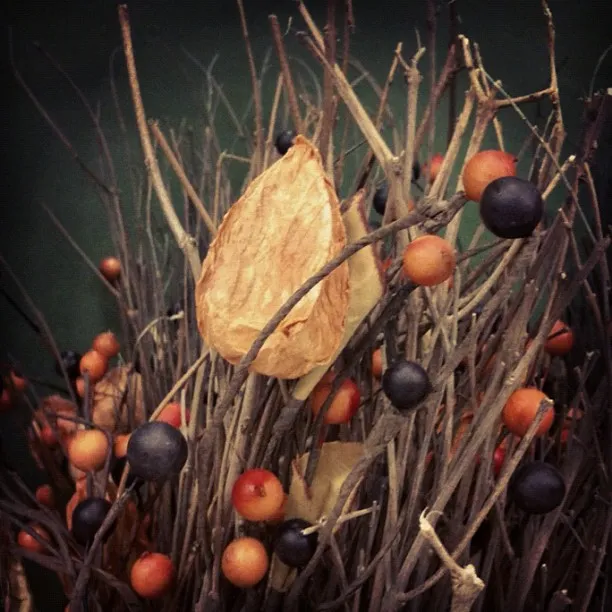 Close-up of a dried floral arrangement with twigs, orange and black berries, and a single papery seed pod in the center.