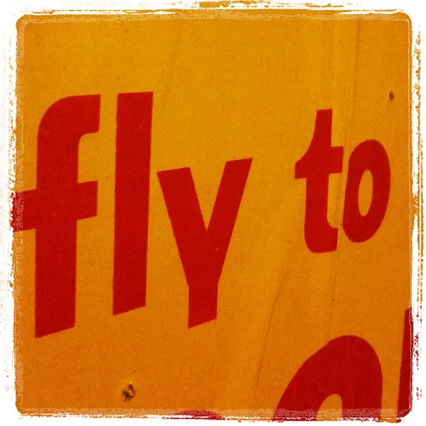 Close-up of a yellow sign with bold red text partially reading “fly to,” showing signs of wear and a distressed border effect.