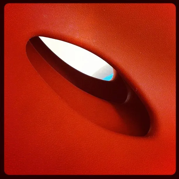 Close-up of a red plastic chair back with an oval cutout, revealing a glimpse of white and blue in the background.