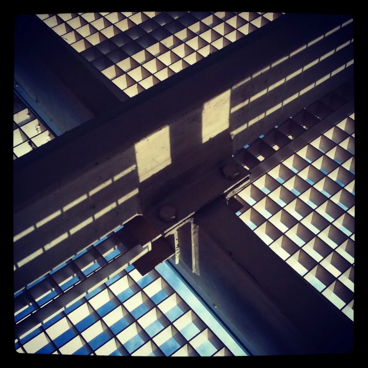 Close-up view looking up at a metal grid structure with sunlight casting sharp geometric shadows on intersecting steel beams.