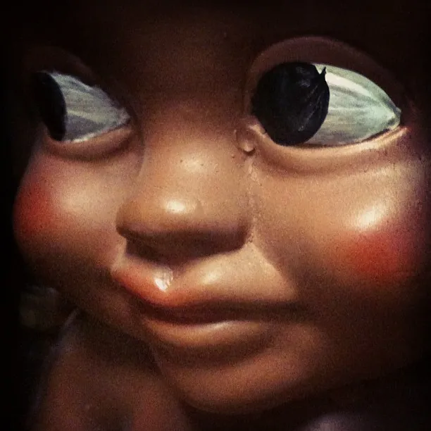 Close-up of a vintage doll’s face with large painted eyes, one of which is chipped, and a visible tear on its cheek.