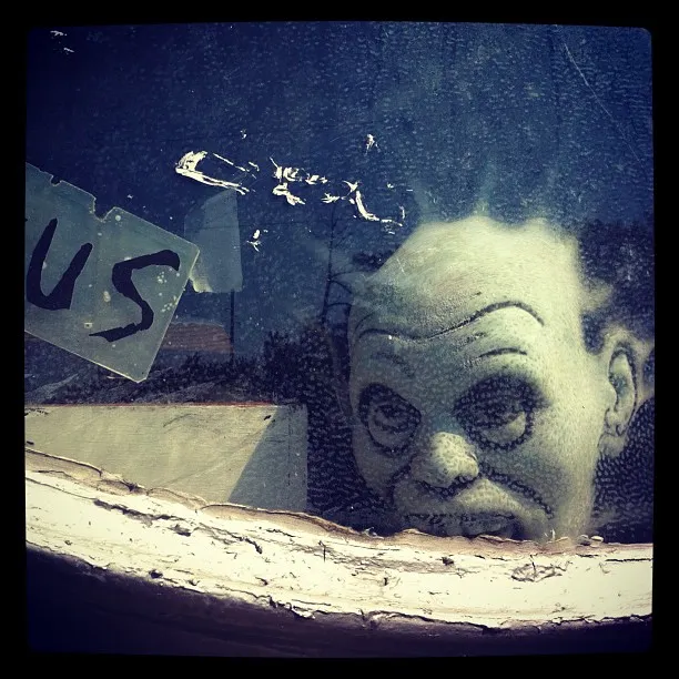Creepy doll or mask of a bald figure with dark eyes peering through a dusty, scratched window with a torn sticker.