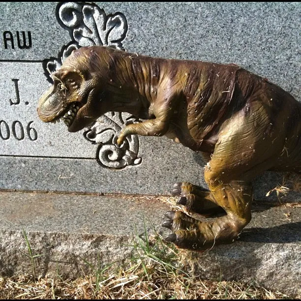 A small plastic Tyrannosaurus rex toy rests against a granite gravestone with decorative carvings and partial text visible.
